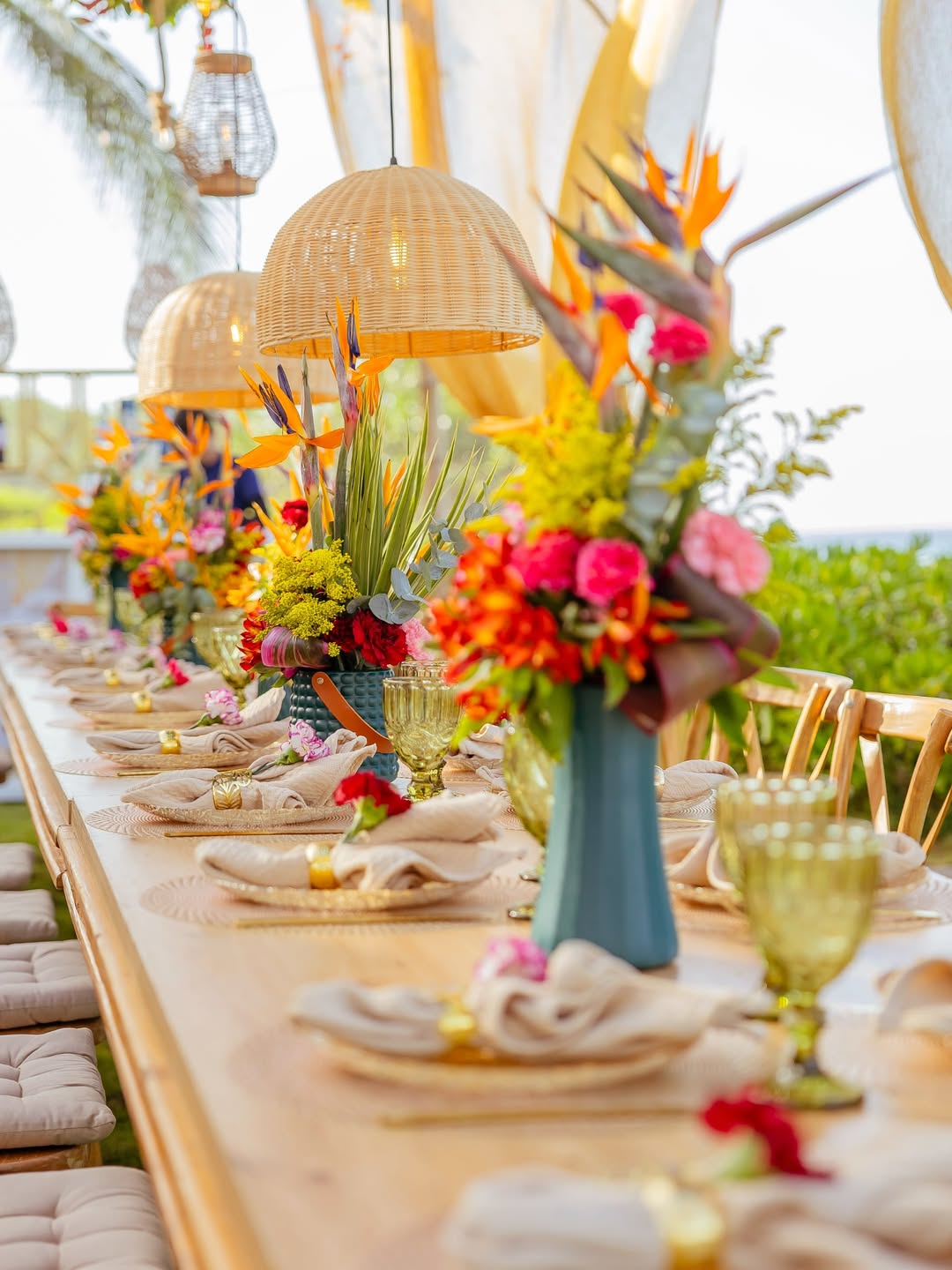 Mesa larga con lámparas de mimbre y centros tropicales para evento al aire libre